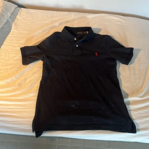 Svart piké från Polo Ralph Lauren - Svart pikétröja från Polo Ralph Lauren med röd broderad logga på bröstet. Modellen är custom slim fit och har klassisk krage, korta ärmar och knappar framtill. Materialet är mjuk bomull som känns skönt mot huden. Perfekt för en clean och stilren look.