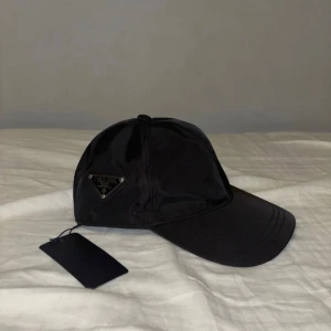 Svart keps i nylon - Svart keps med klassisk böjd skärm och ikonisk metalltriangel på sidan. Tillverkad i glansig nylon som ger en clean och modern vibe. Perfekt accessoar för dig som vill ha en lyxig touch på din outfit.