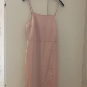 Ljusrosa långklänning från NA-KD - Säljer en stilren ljusrosa långklänning från NA-KD med smala axelband och rak passform. Klänningen har en slits framtill och är tillverkad i ett glansigt satinliknande material som ger en lyxig känsla. Perfekt för dig som gillar minimalistisk och chic stil. Helt oanvänd