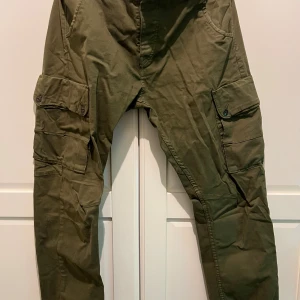 Olivgröna cargopants Jack & Jones - Säljer ett par olivgröna cargopants från Jack & Jones med flera stora fickor på benen och bakfickor med knapp. Byxorna har en loose passform och är tillverkade i bomull, perfekta för en chill och avslappnad stil.