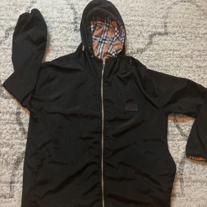 Burberry vändbar vindjacka med huva - Säljer en vändbar Burberry vindjacka med huva. Ena sidan är svart med klassisk logopatch, andra sidan har ikoniskt rutigt mönster i beige, svart, vitt och rött. Jackan har dragkedja, långa ärmar och detaljer vid ärmslut. Perfekt för dig som vill variera din stil. L funkar som M