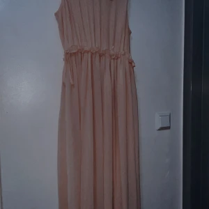 Ljusbeige långklänning från H&M - Superfin långklänning från H&M i en ljusbeige ton. Klänningen har ärmlös design, markerad midja med diskreta volangdetaljer och ett mjukt, plisserat tyg som faller snyggt. Perfekt för dig som gillar en romantisk och stilren look.