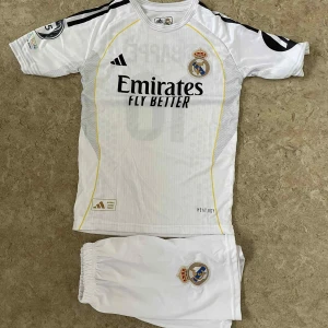 Real Madrid Mbappé fotbollströja Adidas - Säljer en vit Real Madrid fotbollströja från Adidas med Mbappé och nummer 10 på ryggen. Tröjan har korta ärmar, svarta detaljer och gula linjer längs sidorna. Materialet är lätt och andas, perfekt för match eller träning. Real Madrid-emblem på bröstet och shorts med klubbmärke ingår.