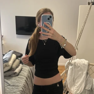 Svart croppad topp från Gina Tricot - Svart croppad topp från Gina Tricot med korta ärmar och snygg snörning på sidorna som ger en rynkad effekt. Toppen har rund hals och är gjord i mjuk bomull, perfekt att styla med höga jeans eller kjol. Köpt för 300kr pris kan diskuteras!