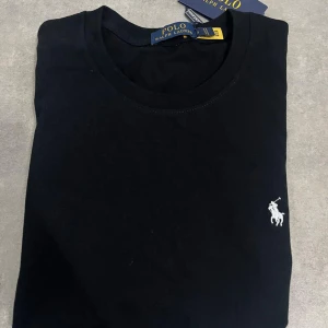 Svart t-shirt från Polo Ralph Lauren - Svart t-shirt från Polo Ralph Lauren med klassisk rund hals och korta ärmar. T-shirten har den ikoniska vita Polo-loggan broderad på bröstet och är tillverkad i mjuk bomull för skön komfort. Perfekt basic med stilren design.