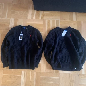 Svart stickad tröja Polo Ralph Lauren - Svart kabelstickad tröja från Polo Ralph Lauren med röd broderad logga på bröstet. Rund hals, ribbade muddar och klassisk passform. Perfekt för dig som gillar stilrena och tidlösa plagg med premiumkänsla.