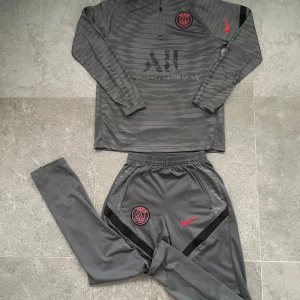 Grå PSG tracksuit Nike  - Säljer en grå långärmad träningströja från Nike och PSG. Tröjan har röd Nike-logga på ärmen och PSG-emblem på bröstet. Materialet är mjukt polyester, perfekt för fotboll eller träning. Snygg och sportig design! Pris kan diskuteras 