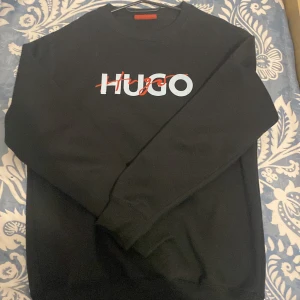 Svart sweatshirt från Hugo Boss - Svart sweatshirt från Hugo Boss med stor HUGO-logga i vitt och rött framtill. Tröjan har rund hals, ribbade muddar och är tillverkad i mjuk bomull. Perfekt för dig som gillar stilrena och snygga märkesplagg.