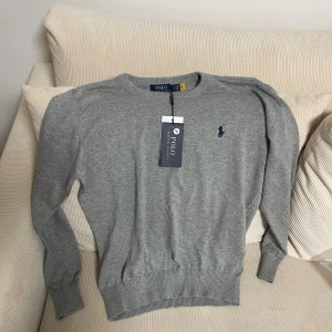 Grå tröja från Polo Ralph Lauren - Snygg grå tröja från Polo Ralph Lauren med klassisk rund hals och ribbade muddar. Tröjan har det ikoniska Polo-logot broderat i mörkblått på bröstet. Perfekt för dig som gillar stilrena och tidlösa plagg.