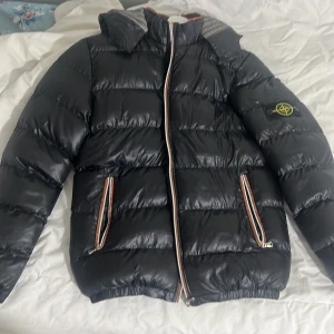 Svart pufferjacka från Stone Island - Svart pufferjacka från Stone Island med ikonisk patch på ärmen. Jackan har huva, dragkedja framtill och två fickor med dragkedja. Insidan är randig i beige, svart och vitt. Perfekt för kalla dagar och har en clean, modern look.