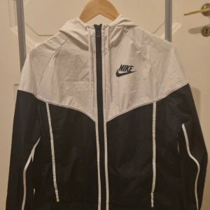 Nike svartvit vindjacka med huva M - Nike vindjacka i svart och vitt med snygg kontrastdesign och logga på bröstet. Jackan har huva, hel dragkedja och vita detaljer längs ärmarna. Perfekt för dig som gillar sportig stil och vill ha något lätt och funktionellt.