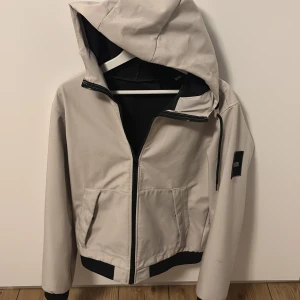 Beige vindjacka från Jack & Jones - Snygg beige vindjacka från Jack & Jones med huva och svarta detaljer. Jackan har dragkedja framtill, två fickor och en svart patch med logga på ärmen. Perfekt för dig som gillar stilren och enkel design.