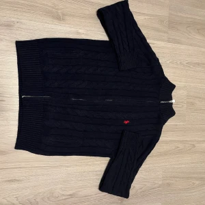 Svart kabelstickad kofta från Polo Ralph Lauren - Svart kabelstickad kofta från Polo Ralph Lauren med röd broderad logga på bröstet. Koftan har långärmad design, ribbade muddar och krage samt dragkedja framtill. Perfekt för dig som gillar klassisk stil med en modern touch. Det står M på tröjan men den har krympt lite så den är som S 