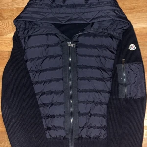 Svart Moncler pufferjacka med huva - Svart pufferjacka från Moncler med stickade ärmar och quiltad front. Jackan har huva, dragkedja framtill och en ficka med dragkedja på ärmen. Moncler-logga på vänster ärm. Materialmix av dun, polyester och stickat ger en schysst look. Storlek L väldigt liten i storlek