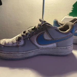 Nike Air Force 1 vit/blå sneakers /ganska slitna  - Nike Air Force 1 sneakers i vit och ljusblå färg med klassisk snörning och perforerad tå. Swoosh-loggan i blått på sidan och blå detalj med Nike Air på hälen. Platt sula och rund tå, tillverkade i skinn och syntetmaterial. Perfekt för dig som gillar retrostil och streetwear.