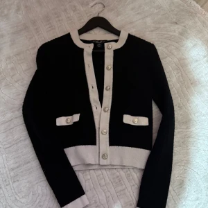 Svart och vit kofta från Amisu XS - Snygg svart och vit kofta från Amisu i stickad material. Koftan har långa ärmar, dekorativa silvriga knappar framtill och vid fickorna, samt kontrastfärgade detaljer vid krage, muddar och nederkant. Perfekt för dig som gillar klassisk och chic stil.