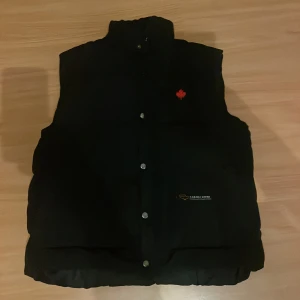 Svart dunväst från Canada Goose - Svart dunväst från Canada Goose med hög krage och tryckknappar framtill. Västen har en liten röd lönnlöv-detalj på bröstet och broderad logotyp på fickan. Klassisk, quiltad design som håller värmen under kyliga dagar.