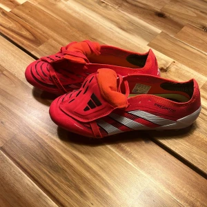 Adidas Predator röda fotbollsskor - Säljer ett par Adidas Predator fotbollsskor i rött med vita och svarta detaljer. Skorna har snörning, låg profil och mönstrad ovandel för extra grepp. Snygg design med klassiska Predator-ränder och text på sidan. Perfekta för dig som vill sticka ut på planen.