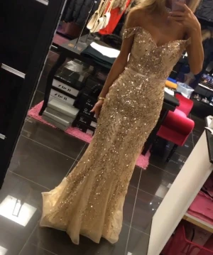 Balklänning - Champagnefärgad långklänning med glittriga paljetter över hela klänningen. Figurnära passform med markerad midja och offshoulder-design. Klänningen har en elegant v-ringning och är tillverkad i ett skimrande syntetmaterial som ger en lyxig känsla.