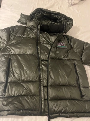 Mörkgrön pufferjacka EA7 Armani - Säljer en snygg mörkgrön pufferjacka från EA7 Emporio Armani. Jackan har en glansig finish, stor huva, två sidofickor med dragkedja och ett tydligt EA7-märke på bröstet. Perfekt för kalla dagar och har en riktigt modern vibe.