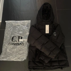 Svart pufferjacka från C.P. Company - Svart pufferjacka från C.P. Company i storlek S. Jackan har huva, dragkedja framtill och två sidofickor med dragkedja. Den är quiltad och har den ikoniska C.P. Company-linsen på ärmen. Perfekt för kalla dagar och har en clean, modern look.