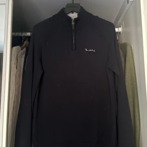 Marinblå halfzip bondelid - Svart stickad tröja från Bondelid med broderad logga på bröstet. Tröjan har en hög krage och half zip-dragkedja framtill. Långärmad och ribbade muddar vid ärmslut och nederkant. Perfekt för kyliga dagar.
