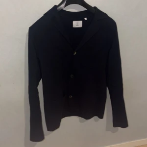 Svart stickad kavaj från Black, M - Svart stickad kavaj från Bläck i storlek M. Jackan har två framfickor, klassisk krage och knäppning med två knappar. Materialet är mjukt och stickat, vilket ger en chill och stilren vibe. Perfekt för dig som gillar enkel och clean look. Butiks pris 1.399kr