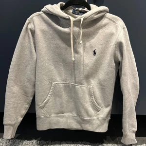 Grå hoodie från Polo Ralph Lauren - Klassisk grå hoodie från Polo Ralph Lauren med broderad marinblå logga på bröstet. Tröjan har huva med snörning, känguruficka och ribbade muddar. Perfekt för chill dagar och enkel att matcha med jeans eller joggers.