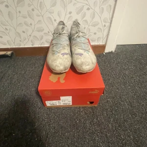 Vita fotbollsskor från Puma - Säljer ett par vita fotbollsskor från Puma med snörning och rund tå. Skorna har en syntetisk ovandel med stickad känsla och färgdetaljer i blått och lila på sidan. Sulan har dobbar för bra grepp på planen. Perfekt för dig som vill ha snygga och funktionella skor till matchen och är på en budget. Ny pris är 1250kr men mitt pris blir 600