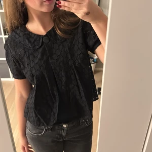 Svart blus med broderade detaljer - Säljer en svart blus med korta puffärmar och rund krage. Blusen har ett broderat hålmönster över hela tyget och stängs med en knapp framtill. Den är luftig och har en söt, feminin look som passar perfekt till jeans eller kjol.