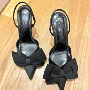 Svarta pumps med rosett från Zara - Använda 2 gånger!! Snygga svarta pumps från Zara med spetsig tå och stor rosett framtill. Skorna har en elegant klack och öppen häl med smal rem runt ankeln. Materialet är textil och syntet, vilket ger en stilren look som sticker ut.