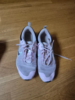 Nike rosa sneakers med snörning - Säljer ett par snygga Nike sneakers i ljusrosa med vita detaljer och svart häl. Skorna har en rund tå, platt sula och är tillverkade i syntetmaterial med mesh-överdel för bra ventilation. Klassisk Nike-logga på sidan och snörning framtill.