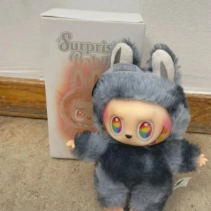Pop Mart Surprise Baby Pretty Series - Unik samlarfigur från Pop Mart Surprise Baby Pretty Series. Dockan har regnbågsfärgade ögon, mjuk grå pälsdräkt med kaninöron och levereras med originalförpackning. Mycket fint skick, inga synliga defekter eller slitage. Perfekt för samlare eller som present.