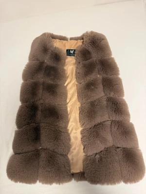 Brun fluffig pälsväst från YXPS - Supermjuk och lyxig brun pälsväst från YXPS med quiltad design. Västen har en varm beige insida och är gjord av täta, fluffiga pälsrutor som ger en cool och trendig look. Perfekt för att höja din outfit under kyliga dagar.