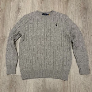 Grå kabelstickad tröja Polo Ralph Lauren - Kabelstickad grå tröja från Polo Ralph Lauren med klassisk rund halsringning och broderad svart logga på bröstet. Tröjan är långärmad och har ribbade muddar vid ärmslut och nederkant. Perfekt för lager-på-lager under kyliga dagar.