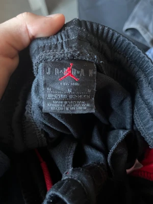 Svarta Jordan mjukisbyxor med röd logga - Snygga svarta mjukisbyxor från Jordan med röda sidopaneler och den klassiska Jumpman-loggan broderad på benet. Byxorna har elastisk midja och muddar vid bensluten, tillverkade i mjukt polyester-material som är perfekt för chill eller träning.