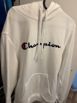 Vit hoodie från Champion, storlek L - Säljer en vit hoodie från Champion i storlek L. Tröjan har broderad Champion-logga i marinblått och rött på bröstet och en mindre logga vid ärmslutet. Klassisk huva med snörning och stor magficka framtill. Mjuk och skön bomullsmix, perfekt för chill dagar.