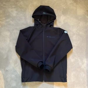 Svart vindjacka från Sail Racing - Svart vindjacka från Sail Racing med huva och hel dragkedja framtill. Jackan har fleecefoder på insidan, ribbade muddar och snygg logga på bröstet samt en rund Sail Racing-detalj på ärmen. Perfekt för blåsiga dagar och sportig stil. Jackan är nästan helt ny. Vid funderingar är det bara att fråga