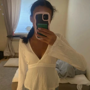 Vit omlottopp med knytband - Säljer en vit omlottopp med långa ärmar och v-ringad hals. Toppen har ett snyggt knytband i midjan som går att justera och ett lätt strukturerat tyg i bomull. Passformen är normal och toppen har en feminin siluett som framhäver midjan. Ny pris 299 säljer för 199.