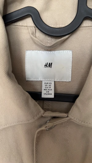 Beige jeansjacka från H&M XS - Snygg beige jeansjacka från H&M i storlek XS. Jackan har klassisk krage, två bröstfickor med knappar och sidofickor. Tillverkad i bomull med silvriga knappar framtill och vid ärmslut. Perfekt för dig som gillar stilrena och enkla plagg.