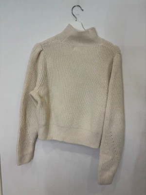 Beige stickad polotröja med håldetaljer - Snygg beige stickad polotröja med lång ärm och ribbad krage. Tröjan har coola håldetaljer längs axlar och ärmar som ger en unik vibe. Modellen är lite croppad och har breda muddar vid nederkant och ärmslut. Perfekt för dig som gillar stilrena och trendiga plagg.