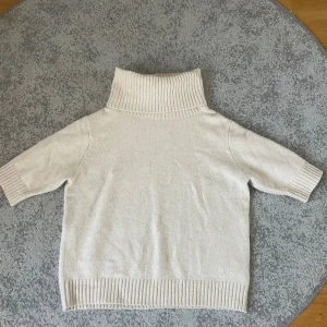 Beige polotröja från Indiska - Säljer en beige polotröja från Indiska med ribbad hög krage och ribbade muddar vid ärmslut och nederkant. Tröjan har korta ärmar och är stickad i mjukt material. Väldigt skön och mysig. Endast använd ett fåtal gånger. 