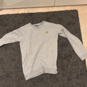 Grå sweatshirt från Lyle & Scott Junior - Snygg grå sweatshirt från Lyle & Scott Junior med klassisk rund hals och långärmade. På bröstet sitter den ikoniska gula fågeln broderad. Perfekt för dig som gillar stilrena och bekväma tröjor med en sportig touch.