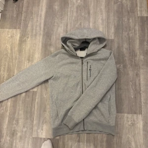 Grå Sail Racing hoodie  - Snygg grå hoodie från Sail Racing med dragkedja framtill och huva. Tröjan har en bröstficka med zip och diskret logga på bröstet. Perfekt för chill eller träning, med ribbade muddar och mjukt material. Tröjan är i storlek M och i mycket bra skick. Nypris ca 1100.