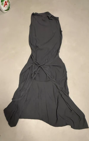 Grå omlottklänning från Ahlvar Gallery - Säljer en lång grå omlottklänning från Ahlvar Gallery i storlek S. Klänningen har ett snyggt fall och är tillverkad i en mjuk blandning av viskos och linne. Den har knytband i midjan och stilren söm längs ryggen för extra detalj. Perfekt för dig som gillar minimalistisk stil.