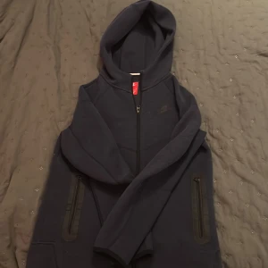 Mörkblå Nike hoodie med dragkedja - Säljer en mörkblå hoodie från Nike med hel dragkedja framtill och två fickor med dragkedja. Tröjan har huva och är tillverkad i mjukt material, perfekt för chill eller träning. Nike-logga på bröstet ger en clean och sportig look.