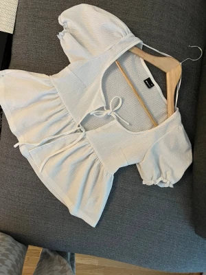 Vit blus med puffärm från Gina Tricot - Supersöt vit blus från Gina Tricot i storlek XS. Blusen har puffiga korta ärmar, omlottkänsla med knyt framtill och volang nedtill. Materialet är lätt strukturerat och känns luftigt, perfekt för varmare dagar. Snygg att styla med jeans eller kjol!