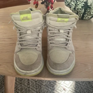 Nike Air Jordan 1 i beige och lime - Nike Air Jordan 1 sneakers i beige mocka och canvas med limegröna detaljer på swoosh, plös och innersula. Klassisk high-top siluett med snörning och rund tå. Perfekta för dig som gillar streetwear och vill sticka ut med en unik färgkombo.
