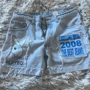 Levi's ljusblå denim shorts med tryck - Unika ljusblå denim shorts från Levi's 550 med relaxed fit. Framsidan har råa kanter och två stora tryck: ett med Playboy-loggan och ett med blå text 'EXCELLENT 2008 THE BEST JEANS'. Klassiska fem fickor och Levi's läderpatch bak. Perfekt för dig som gillar statement-plagg.