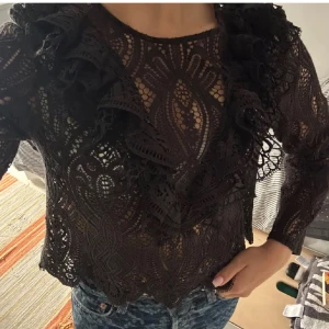 Svart spetsblus från Zara - Supersnygg svart spetsblus från Zara med långa ärmar och volangdetaljer framtill. Blusen har ett genombrutet spetsmönster och rund halsringning. Perfekt att styla med jeans eller kjol för en trendig look.